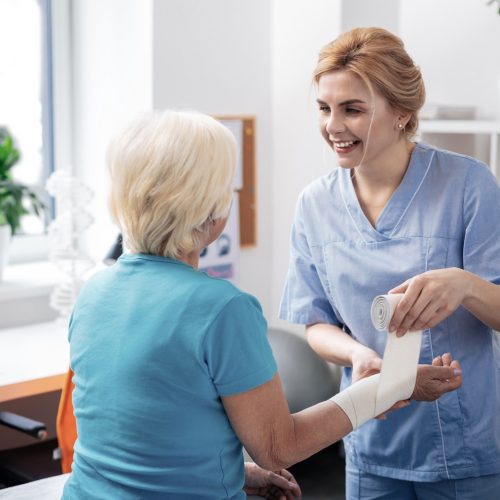 Joyful positive nurse wrapping bandage around her patients hand